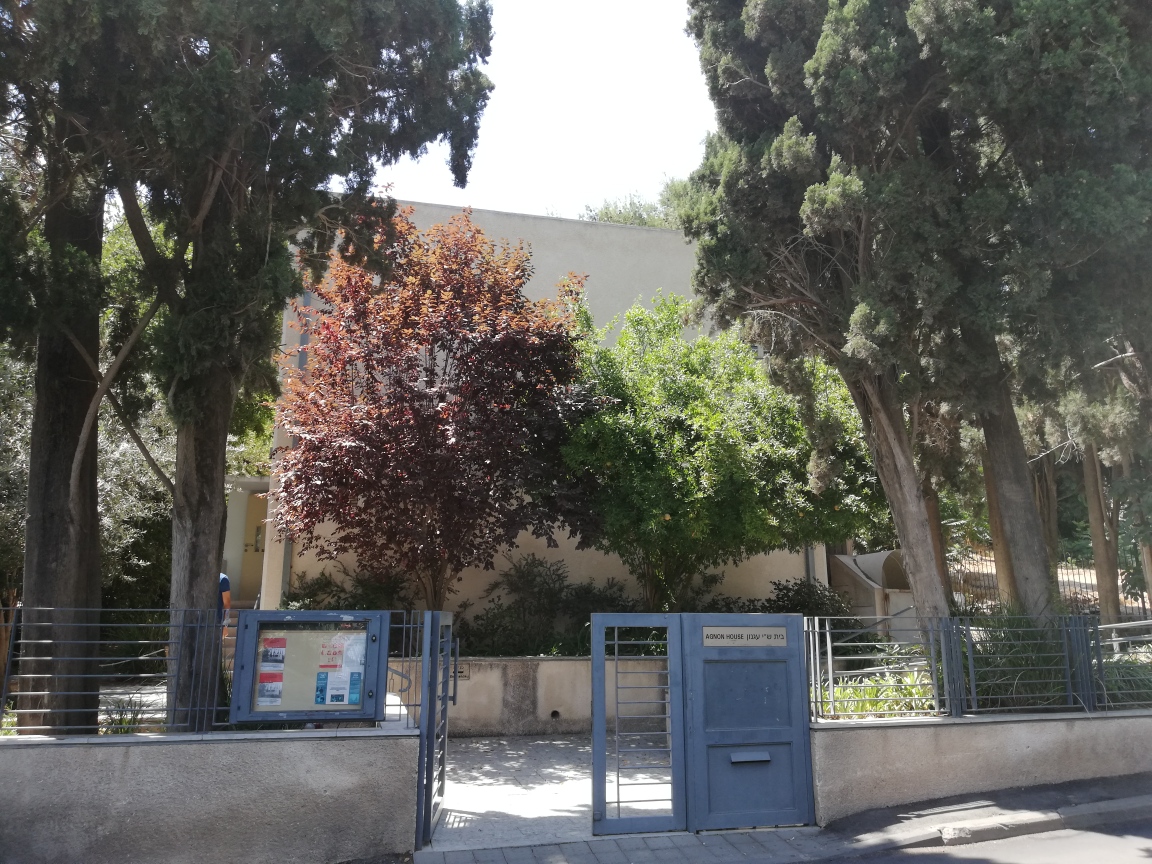 חזית מבנה ציבורי נמוך מאחורי עצים ושער כניסה..  בית עגנון